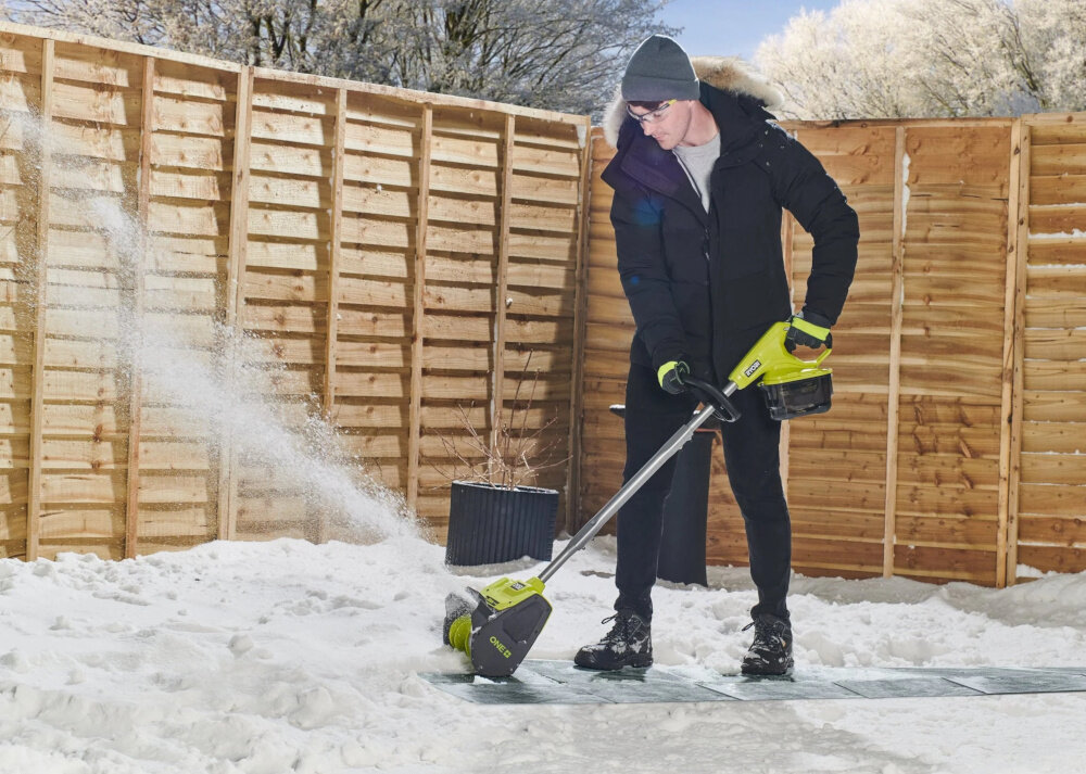 Odśnieżarka akumulatorowa RYOBI RY18ST25A-140 podwórko dom osoba odśnieża taras przed domem w tle drewniana ściana płotu ośnieżone drzewa zasięg wyrzutu śniegu do 6 metrów skutecznie odprowadza śnieg poza oczyszczaną powierzchnię usprawnia całą pracę i poprawia jej efektywność zachowaniu porządku oraz estetyki wokół domu