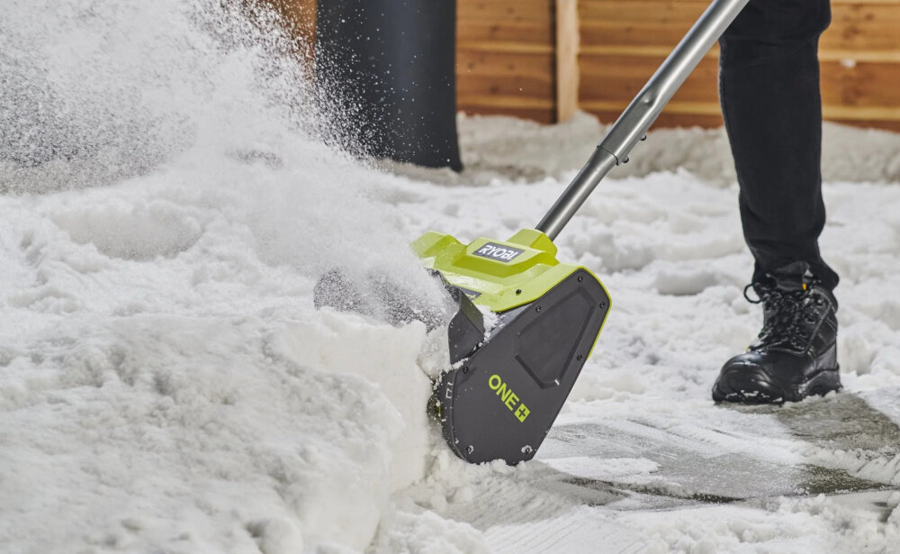 Odśnieżarka akumulatorowa RYOBI RY18ST25A-140 podwórko śnieg osoba odśnieża chodnik w tle drewniana ściana domu płynne przesuwanie po nawierzchni bez konieczności ręcznego odgarniania śniegu zmniejsza to zmęczenie oraz przeciążenie mięśni szybciej oczyścić większą powierzchnię bez przerw