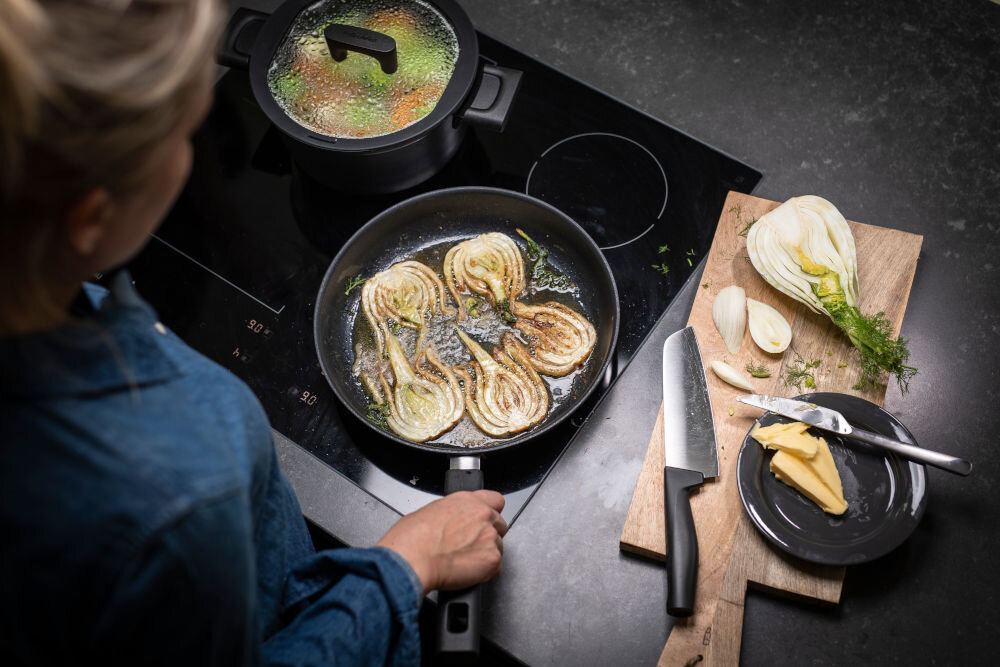 Nóż FISKARS Hard Edge 1051761 osoba smaży danie na patelni obok leży nóż na desce z czarną rączką Ostrze z wysokiej jakości japońskiej stali nierdzewnej precyzja efektywność trwałość dynamiczne cięcie z dużą siłą subtelna precyzja w codziennym gotowaniu przy wymagających technikach kulinarnych