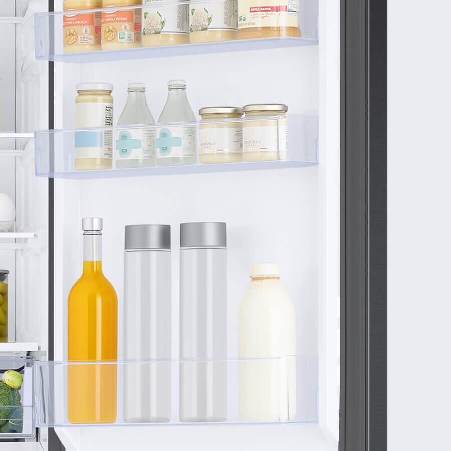 Wnętrze lodówki z widocznymi półkami na drzwiach, na których znajdują się różne butelki i słoiki z napojami oraz produktami spożywczymi. Na górnej półce widoczne są słoiki z przetworami i butelki z napojami, a na dolnej większe butelki z napojami.