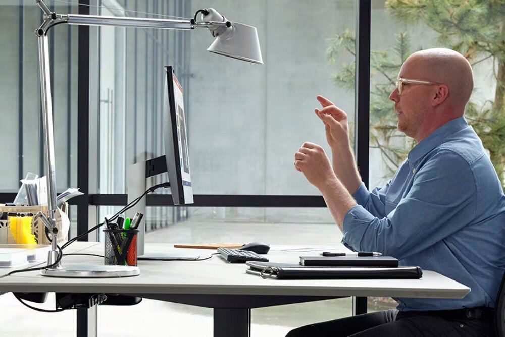 Komputer HP ProOne 440 G9 Mężczyzna siedzi przy biurku w jasnym biurze, patrząc na ekran komputera i gestykulując rękami, obok znajduje się lampka biurkowa i przybory do pisania, opis