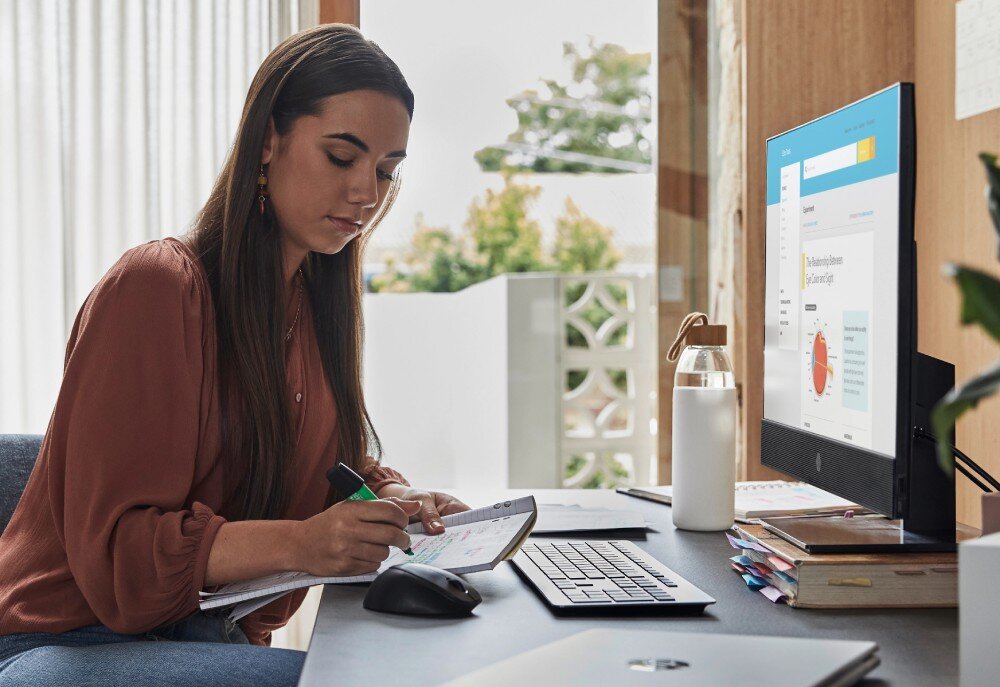 Komputer HP ProOne 440 G9 Kobieta siedzi przy biurku w domowym biurze, pisze w notesie i patrzy na ekran komputera, obok leży myszka, klawiatura i butelka z wodą, obudowa, ekran, matryca
