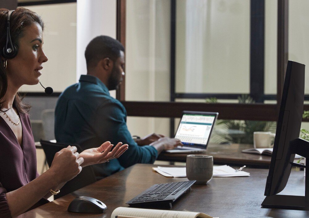 Komputer HP ProOne 440 G9 Kobieta w zestawie słuchawkowym siedzi przy biurku i rozmawia online, za nią mężczyzna pracuje na laptopie, oboje w jasnym biurze, kamera internetowa, mikrofon, głośniki