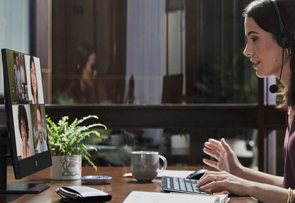 Komputer HP ProOne 440 G9 Kobieta w słuchawkach prowadzi wideokonferencję, na ekranie widać cztery osoby, obok na biurku stoi kubek i doniczka z rośliną, zabezpieczenia ochrona