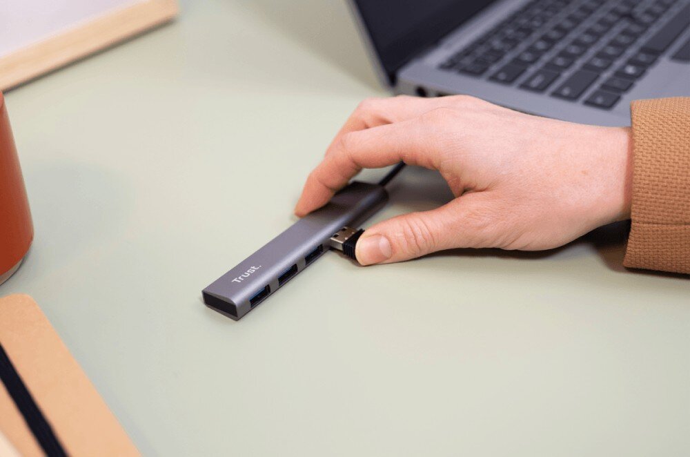 Hub TRUST Halyx 24948 Dłoń w beżowym swetrze wpinająca przewód do portu USB laptopa leżącego na jasnozielonym biurku z notesem i kaktusem w tle
