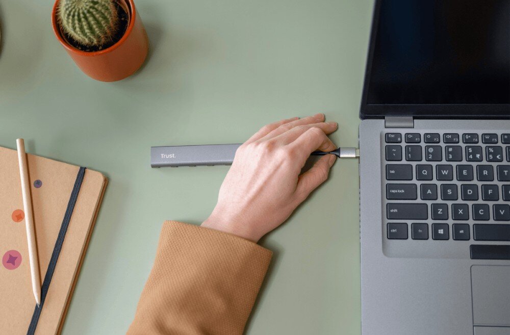 Hub TRUST Halyx 24948 Zbliżenie na rękę podłączającą pendrive do szarego rozdzielacza USB leżącego na białej podkładce biurkowej