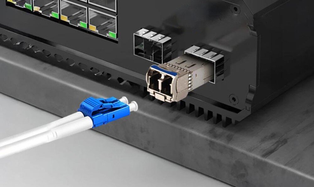 Moduł SFP+ EXTRALINK SFP+ 10G 2-pack Moduł SFP w trakcie podłączania do przełącznika sieciowego, obok leży odłączony kabel światłowodowy z niebieską wtyczką