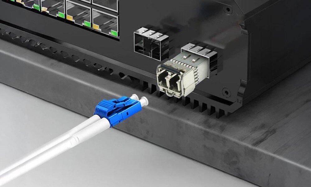 Moduł SFP+ EXTRALINK 10G 2-pack LC/UPC 850nm 300m HP/Aruba Moduł SFP wsunięty do portu w czarnym urządzeniu sieciowym i leżący przed nim kabel światłowodowy z niebieskim złączem LC