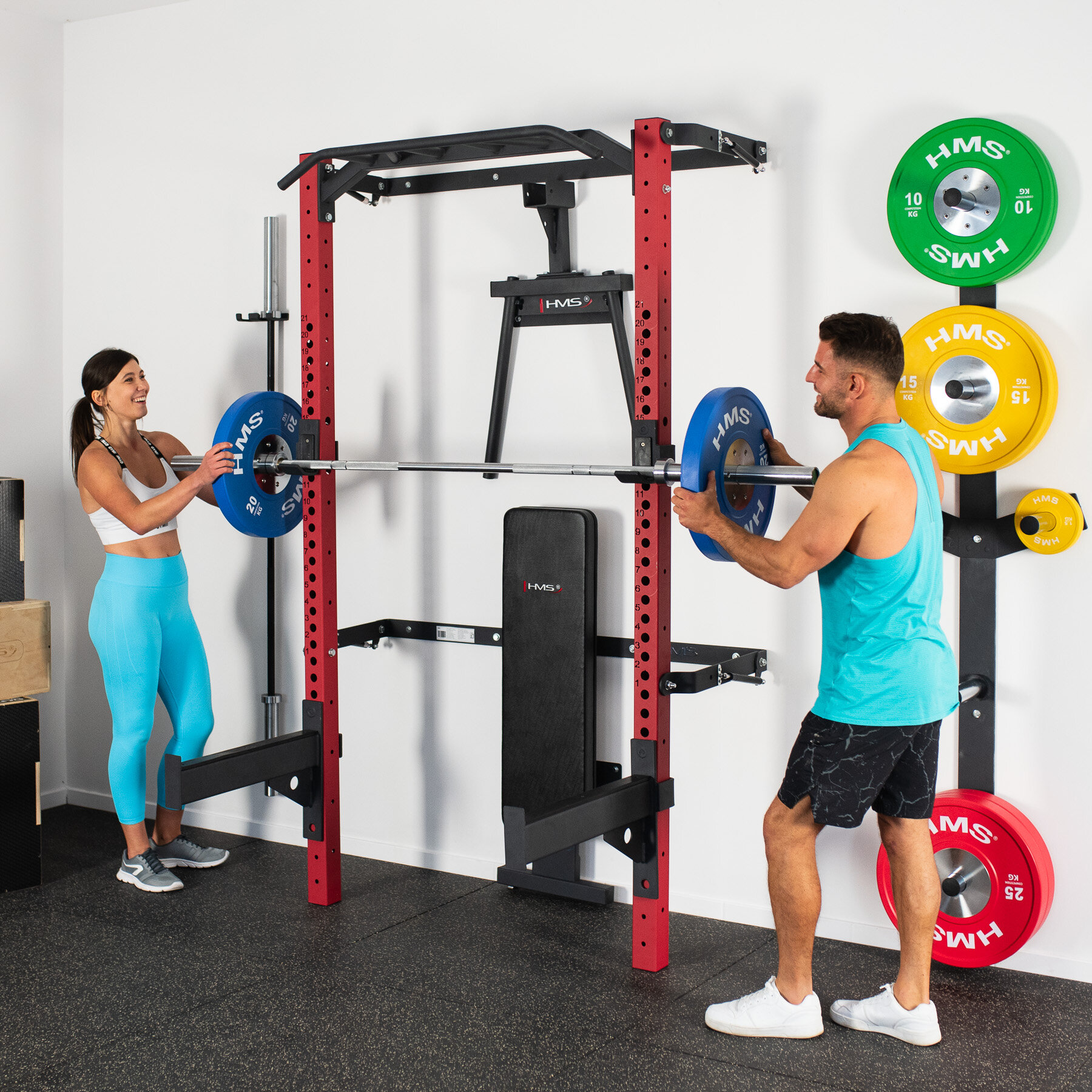 Klatka treningowa HMS KLT22 Czerwony do ściany jest zamontowana klatka ławeczka do ćwiczeń jest złożona do ściany na stojaku prezentują się talerze, kolorowe bumpery a po przeciwnym boku klatki jest uchwyt na którym są zaprezentowane dwa gryfy przy klatce stoi para i zakłada talerze na gryf domowa siłownia składana klatka trening w domu