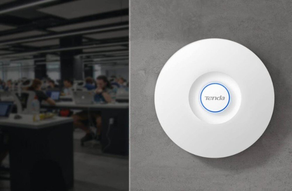 Punkt dostępu TENDA i29 Okrągłe białe urządzenie z niebieskim pierścieniem i logo marki zamontowane na szarej ścianie, w tle rozmyty widok biura typu open space z pracownikami przy biurkach