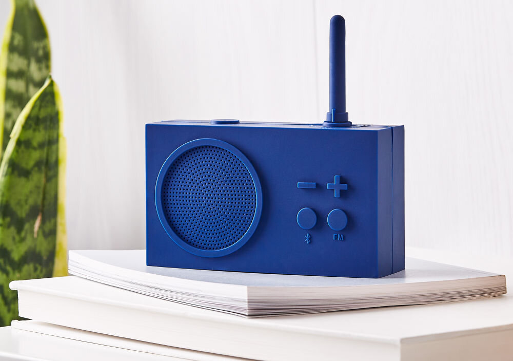 Niebieski radioodtwarzacz z widocznym głośnikiem, anteną oraz przyciskami oznaczonymi symbolami \'-\', \'+\', Bluetooth i \'FM\'. Urządzenie stoi na stosie książek na jasnym tle.