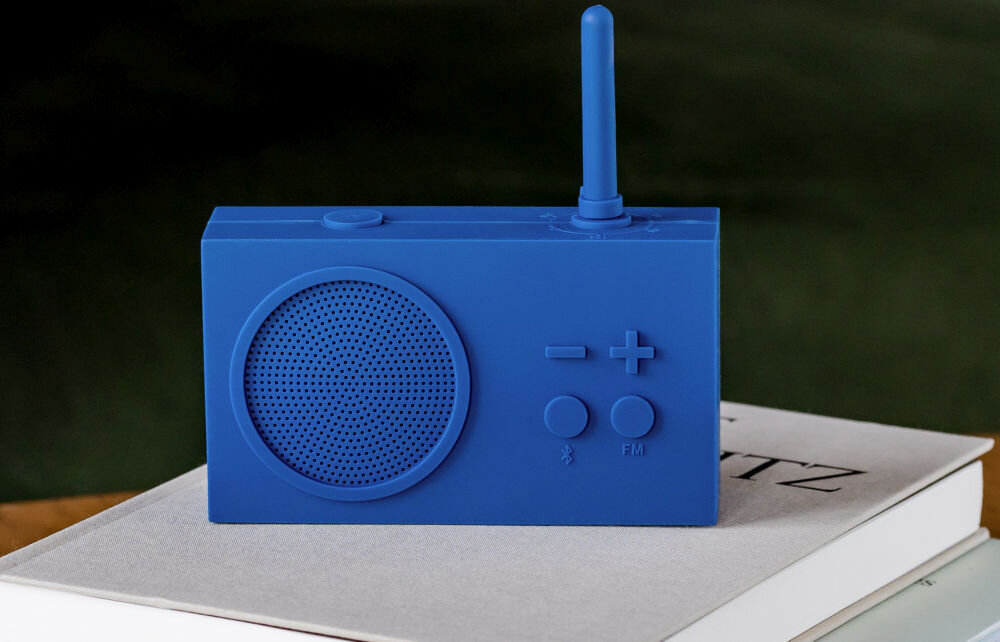 Niebieski radioodtwarzacz z anteną, dużym głośnikiem i przyciskami oznaczonymi symbolami \'-\', \'+\', Bluetooth i FM. Urządzenie stoi na książce z widocznym fragmentem tekstu \'TZ\'.
