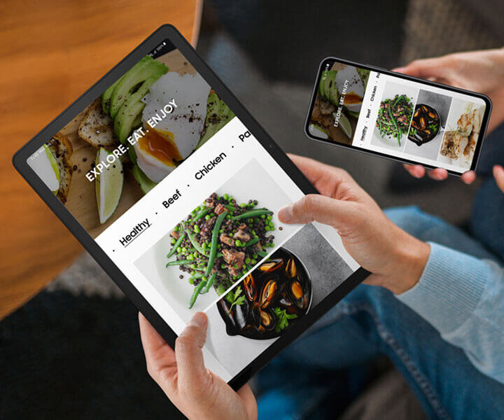 Dwie osoby siedzą obok siebie. Osoba po lewej trzyma Galaxy Tab A9 Plus, a osoba po prawej trzyma smartfon Galaxy. Oba urządzenia wyświetlają tę samą stronę internetową o jedzeniu.