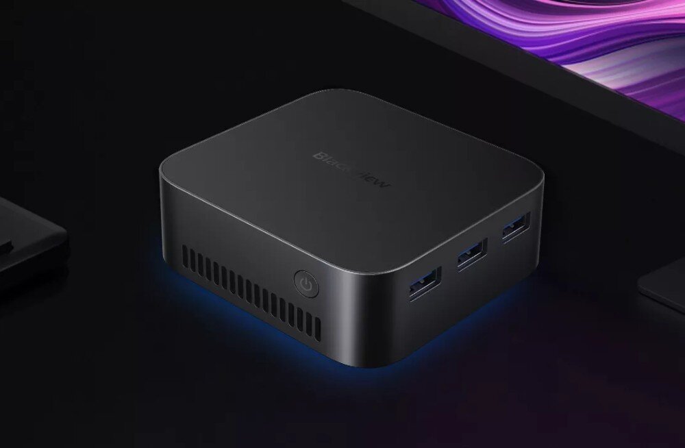 Komputer BLACKVIEW MP80 Małe czarne urządzenie o zaokrąglonych krawędziach z trzema portami USB na bocznej ściance i przyciskiem zasilania z przodu, stojące na ciemnym biurku obok monitora i klawiatury, opis