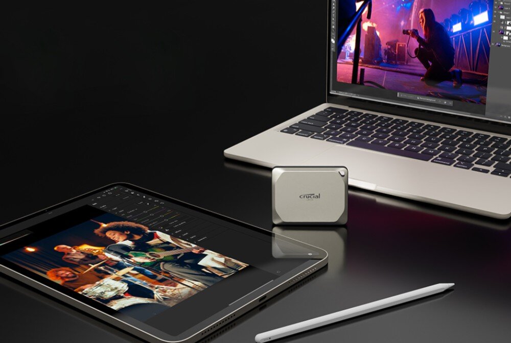 Dysk CRUCIAL X9 Pro for Mac Na ciemnym biurku leżą tablet z wyświetlonym zdjęciem muzyków, laptop z otwartym edytorem graficznym, przenośny dysk w kolorze srebrnym oraz rysik, pojemność, prędkość