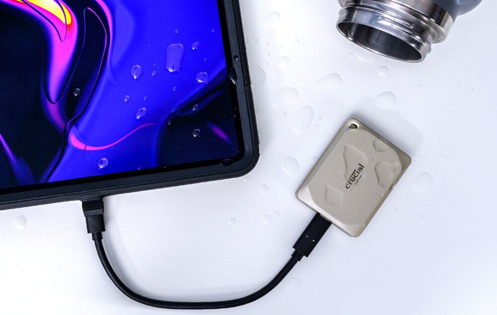 Dysk CRUCIAL X9 Pro for Mac Dysk w kolorze srebrnym leży na białej powierzchni obok tabletu z fioletowo-niebieskim ekranem, wokół widoczne są krople wody, odporność, budowa