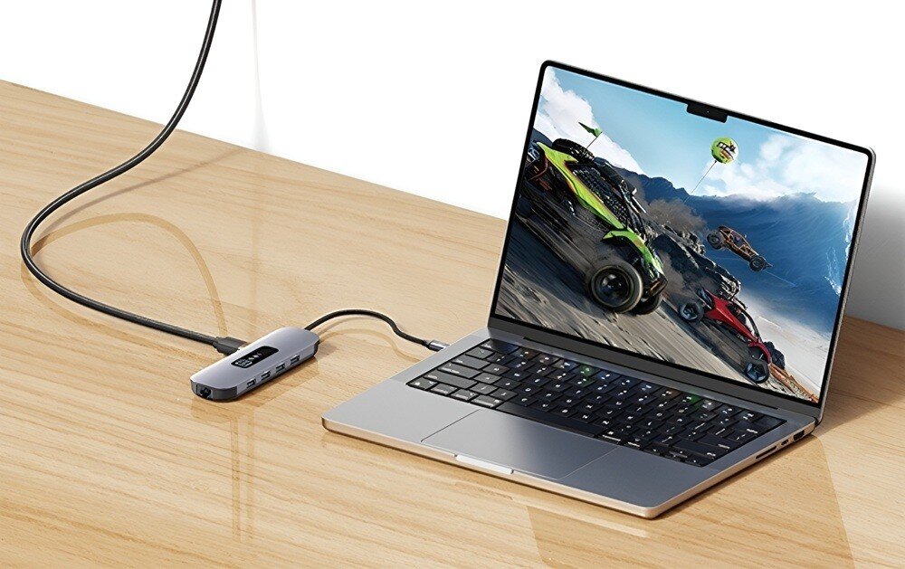 Hub USAMS SJ644HUB01 Laptop stoi na drewnianym biurku. Do portu USB-C podłączony jest hub, a na ekranie widoczna jest gra wyścigowa z samochodami terenowymi, 
            hub adapter technologia gadżet komputer laptop