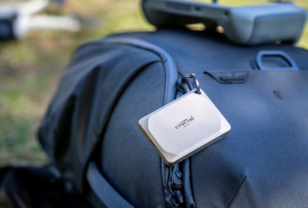Dysk CRUCIAL X9 Pro for Mac Mały dysk w jasnej obudowie przymocowany jest do plecaka za pomocą karabińczyka, w tle widać rozmytą zieloną trawę i część sprzętu elektronicznego, materiał, wygląd