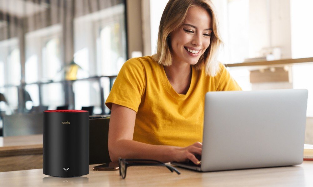 Router CUDY M3000 Kobieta w żółtej koszulce siedzi przy stole i pracuje na laptopie. Obok niej stoi cylindryczne urządzenie sieciowe z logo 'cudy', opis