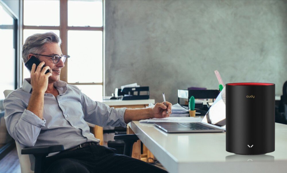 Router CUDY M3000 Mężczyzna w biurze siedzi przy biurku, rozmawia przez telefon i notuje coś na kartce. Na biurku obok laptopa znajduje się cylindryczne urządzenie sieciowe 'cudy', konfiguracja, interfejs, kontrola rodzicielska
