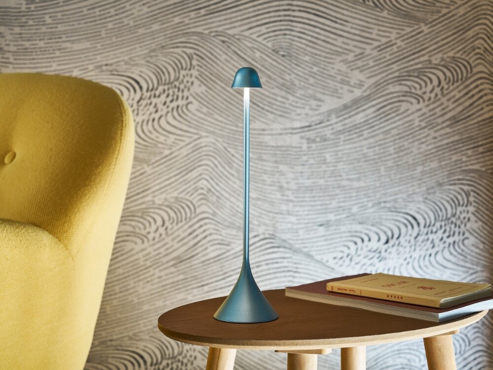 Lampa stołowa LEXON Steli Bell LH95B-LB1 Jasnoniebieski niebieska lampka stoi na stoliku obok książek styl funkcjonalność dotykowe sterowanie możliwość regulacji jasności wodoodporność na poziomie IPX4 w wielu zastosowaniach na stolik nocny taras stół w jadalni do 10 godzin pracy na jednym ładowaniu ładowanie około 3 godziny stylowy design wszechstronność w pomieszczeniach na zewnątrz  