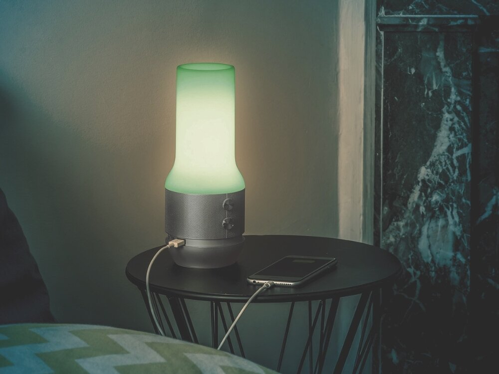 Lampka stołowa LEXON Terrace+ LA134D z głośnikiem i powerbankiem Złoty lampka stoi na stoliku szara obudowa przyciski lampa świeci na kolor zielony obok ładuje się telefon przewód podłączony do lampy powerbank o pojemności 4000 mAh dwa porty USB-A ładowanie dwóch urządzeń jednocześnie podczas aktywności na świeżym powietrzu obudowa z certyfikatem IPX4 odporna na zachlapania w różnych warunkach atmosferycznych funkcjonalność styl do każdej sytuacji 