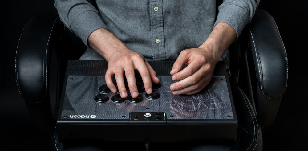 Kontroler NACON Daija Arcade Stick prezentacja kontrolera w codziennym użytkowaniu mężczyzna siedzący na fotelu i korzystający z kontrolera na ciemnym tle joystick i przyciski SANWA wybrane przez zawodowych graczy wysoka precyzja błyskawiczna reakcja trwałość wsparcie w dynamicznej walce kontrola nad ruchami płynność