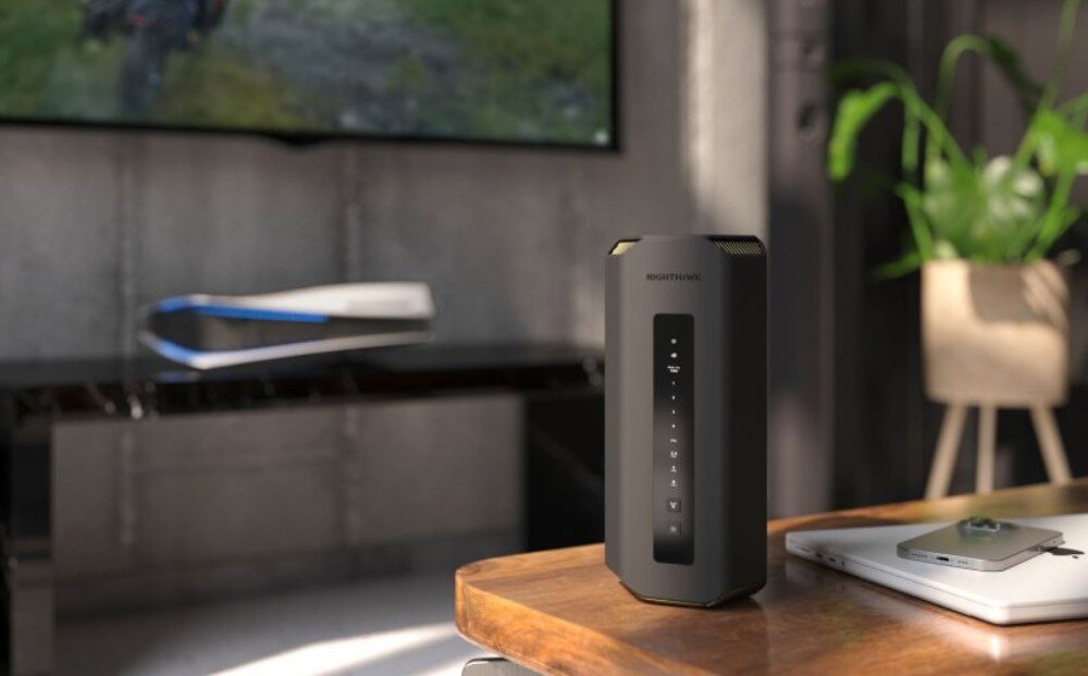 Router NETGEAR Nighthawk RS700S Pionowe urządzenie sieciowe o przekroju graniastosłupa stoi na drewnianym stole obok laptopa i telefonu, w tle widać rozmazany salon z telewizorem, opis