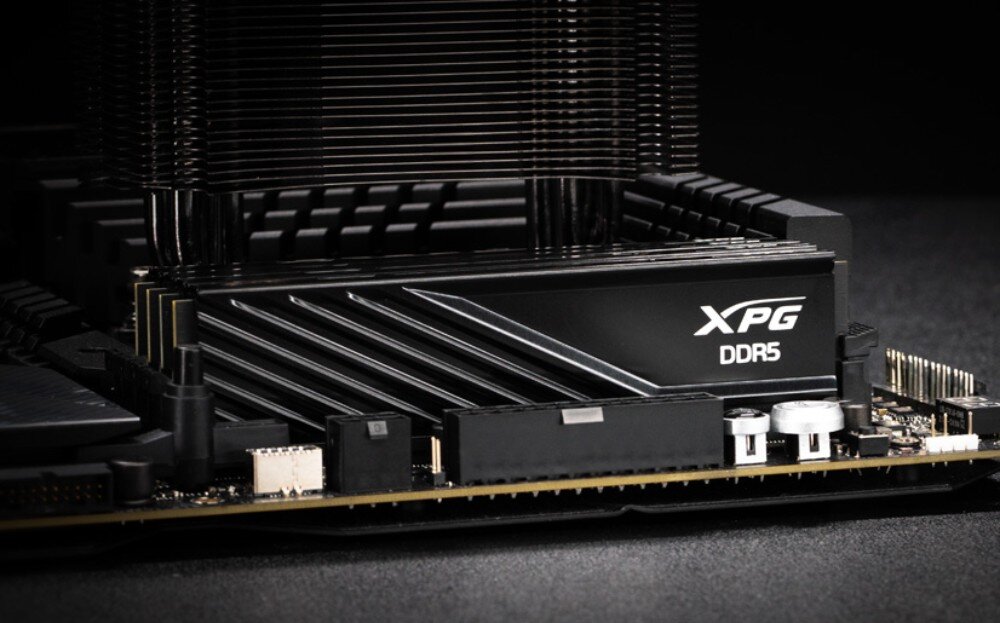 Pamięć RAM ADATA XPG Lancer Blade Zbliżenie na czarny moduł pamięci zainstalowany w gnieździe płyty głównej bezpośrednio pod aluminiowymi żeberkami chłodzenia procesora, widoczne jest logo i napis 'DDR5' na boku obudowy, radiator, wielkość