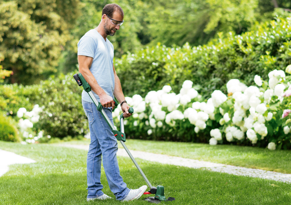 Podkaszarka akumulatorowa BOSCH UniversalGrassCut 06008C1D05 ogród trawnik osoba przycina trawę w tle kwiaty krzewy zadbany trawnik swoboda pracy elastyczność