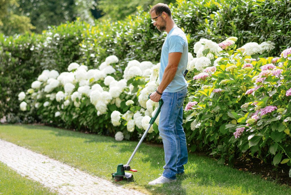 Podkaszarka akumulatorowa BOSCH UniversalGrassCut 06008C1D05 ogród trawnik osoba przycina trawę w tle kwiaty krzewy przycięcie krawędzi nawet ośmiu kortów tenisowych przy użyciu jednego akumulatora o pojemności 2,5 Ah wyraźnie większa efektywność pracy czas ładowania około 2,1 godziny dwa akumulatory o pojemności 2,5 Ah ładowarka