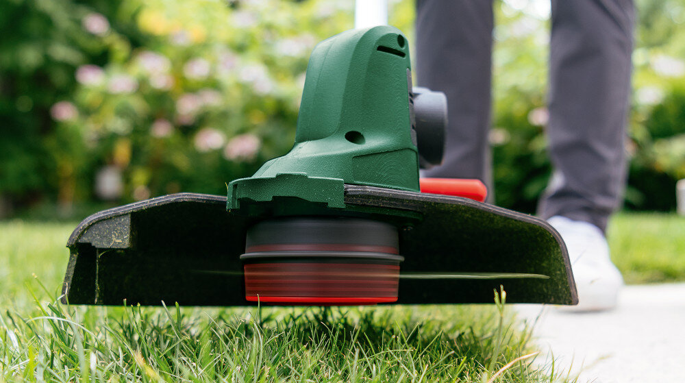 Podkaszarka akumulatorowa BOSCH UniversalGrassCut 06008C1D05 trawnik zbliżenie na głowicę tnącą ogród trawnik osoba przycina trawę w tle kwiaty krzewy głowicę można przechylać bez użycia rąk podcinanie trawy wzdłuż ścieżek, pod ławkami czy przy krawędziach rabat łatwe manewrowanie urządzeniem w trudno dostępnych miejscach prędkość obrotowa do 7300 obr./min żyłka o długości 6 metrów na szpuli