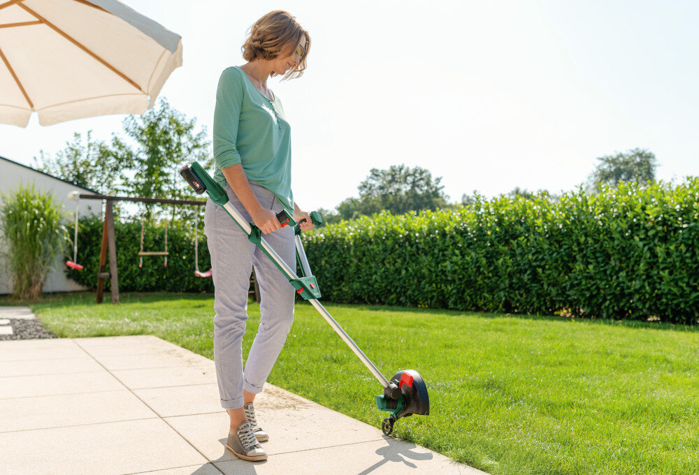 Podkaszarka akumulatorowa BOSCH UniversalGrassCut 06008C1D05 ogród trawnik osoba przycina trawę w tle kwiaty krzewy prosta zmiana z koszenia na przycinanie krawędzi za pomocą jednego przycisku przyspiesza pracę eliminuje konieczność używania osobnych narzędzi do wykańczania krawędzi oszczędność czasu i duży komfort pracy elastyczność