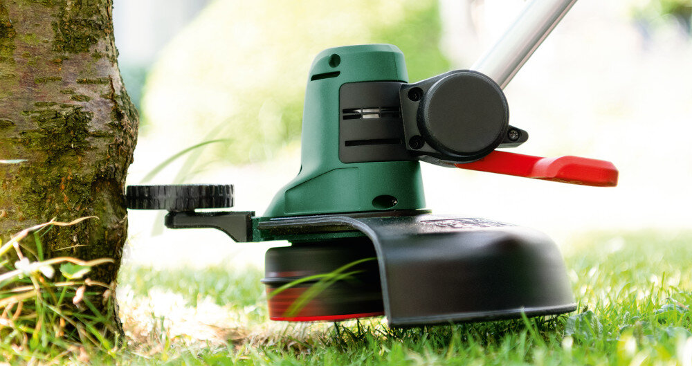 Podkaszarka akumulatorowa BOSCH UniversalGrassCut 06008C1D05 trawnik drzewo zbliżenie na głowicę tnącą cięcie wokół drzewa praktyczny kabłąk dystansowy chroni delikatne rośliny przed przypadkowym uszkodzeniem podczas pracy użytkownik może z dużą precyzją manewrować narzędziem