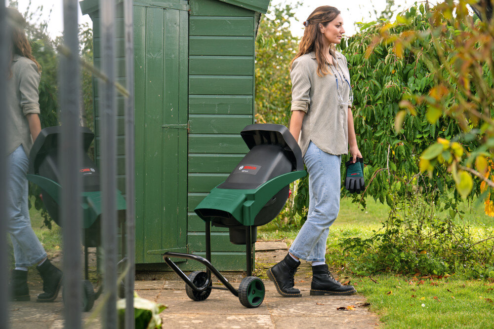 Rozdrabniacz do gałęzi BOSCH AXT Rapid 2000 0600853501 ogród szopa garaż osoba ciągnie za sobą rozdrabniacz w tle drzewa krzewy waga około 11,9 kg dwa kółka uchwyt transportowy wygoda mobilność