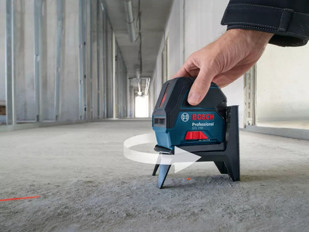 Osoba trzyma laser liniowy BOSCH Professional GCL 2-50, umieszczony na podłodze w korytarzu budynku w trakcie budowy. Na urządzeniu widoczne jest logo BOSCH oraz napis \'Professional GCL 2-50\'.