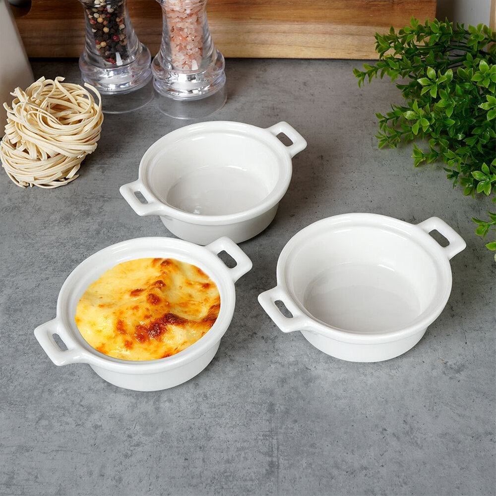 Na obrazie znajdują się trzy białe porcelanowe miseczki do zapiekania, z których jedna zawiera zapieczoną potrawę. W tle widoczne są przyprawy, makaron i zielona roślina.