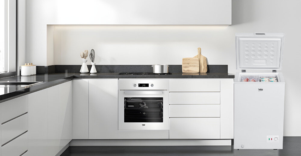 ZAMRAŻARKA BEKO CF100EWN Jasna, minimalistyczna kuchnia z białą zabudową i ciemnym blatem, w której po prawej stronie stoi otwarta zamrażarka skrzyniowa Beko ustawiona na równi z linią szafek