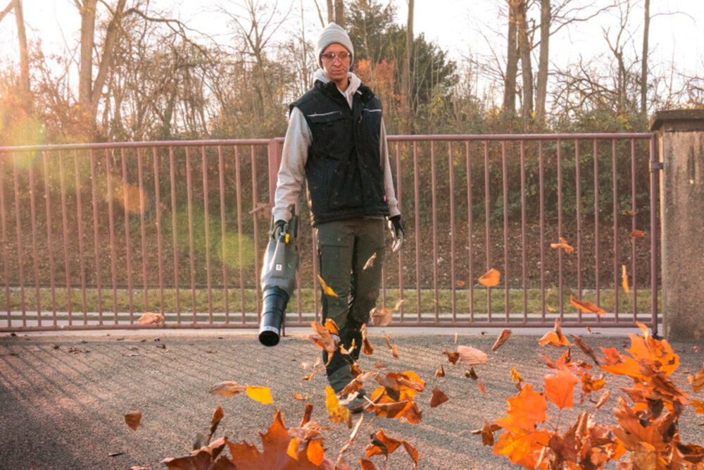 Dmuchawa do liści akumulatorowa KARCHER LB 930-36 Bp Pack 1.042-508.0 ogród podjazd osoba zdmuchuje liście z podjazdu w tle metalowy płot drzewa krzewy niebo zrównoważona konstrukcja ergonomiczna pozycja podczas pracy długie użytkowanie bez obciążania pleców czy ramion waga 3 kg