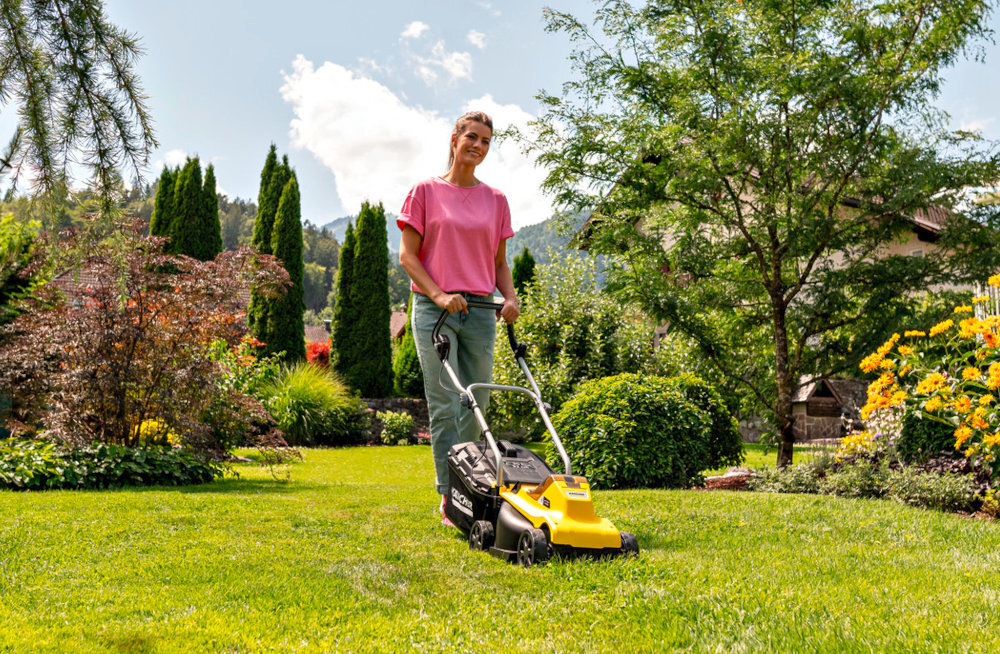 Kosiarka akumulatorowa KARCHER LMO 2-18 1.445-400.0 ogród trawnik osoba kosi trawnik w tle drzewa krzewy kwiaty niebo obok dom zadbany ogród efektywne koszenie mobilność