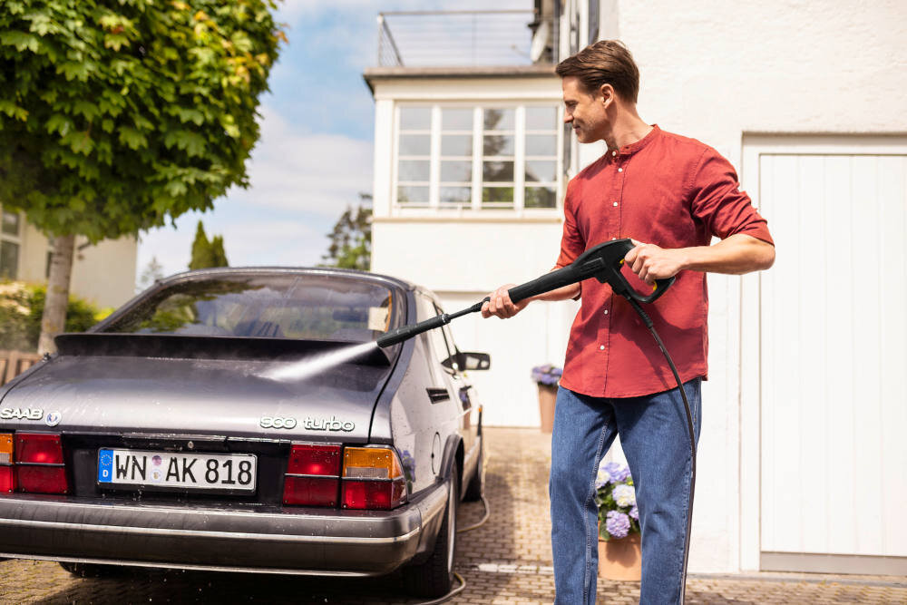 Osoba używa myjki ciśnieniowej do mycia samochodu marki Saab 900 turbo. Na tablicy rejestracyjnej widnieje napis \'WN AK 818\'.