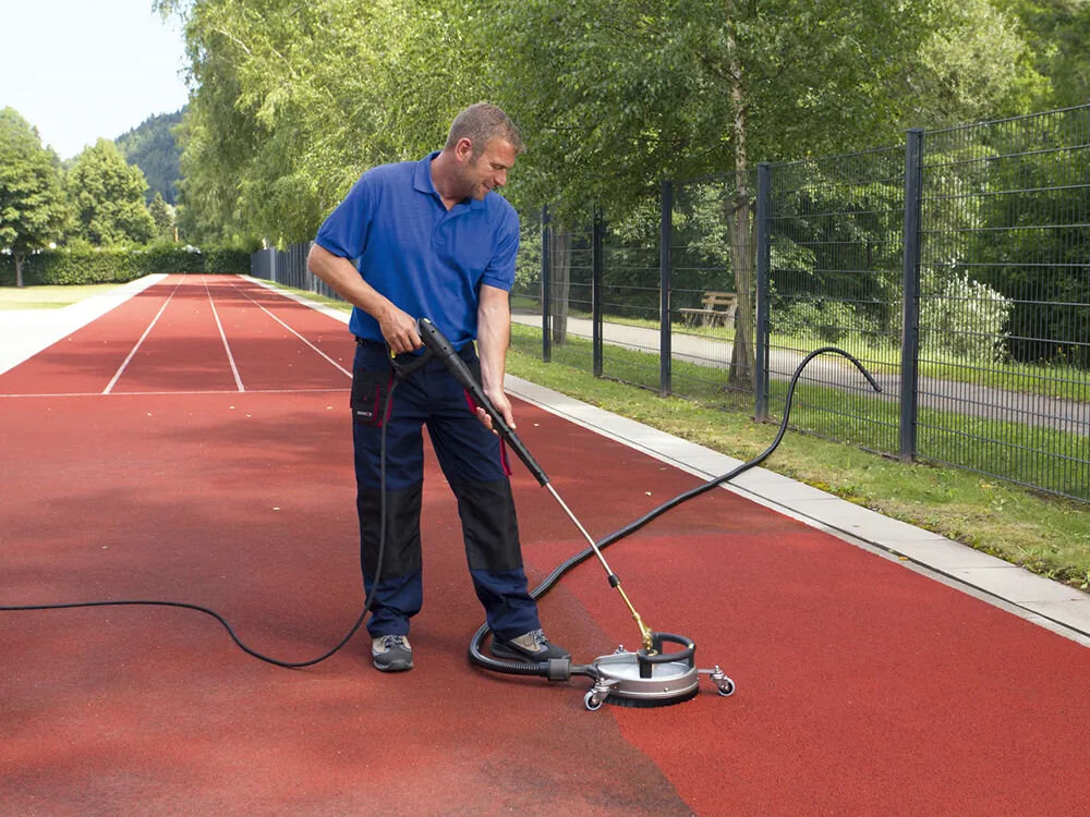 Szczotka KARCHER FRV 30 ME 2.642-911.0 na czerwonej bieżni stoi mężczyzna, który czyści ją za pomocą myjki ciśnieniowej z okrągłą szczotką, usuwając brud i zanieczyszczenia