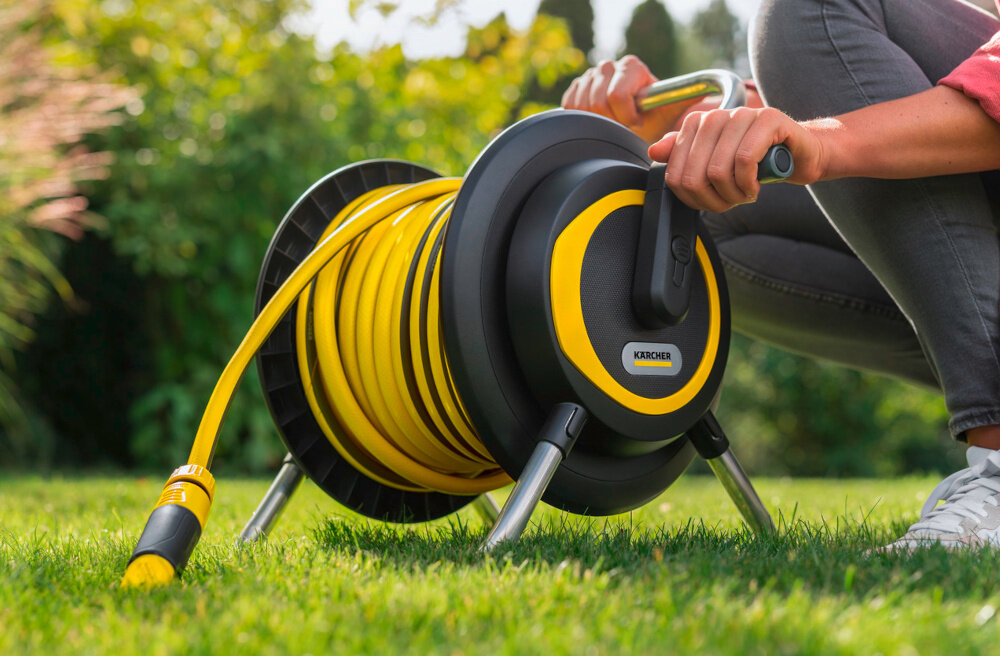 Bęben na wąż KARCHER Premium HR 4 2.645-373.0 ogród trawnik bęben z nawiniętym wężem ogrodowym stoi na trawie obok klęczy osoba trzyma dłoń na korbie w tle drzewa porządek wygodne podlewanie przechowywanie węża ogrodowego