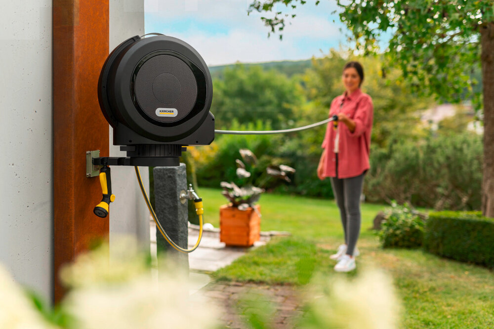 Bęben na wąż KARCHER HBX 5.30 Automatyczny 2.645-380.0 ogród bęben umieszczony na ścianie budynku w tle osoba wyciąga wąż ogrodowy z bębna dalej drzewa kwiaty niebo wygodne przechowywanie węża praktyczne zastosowanie przechowywanie węża ogrodowego podlewanie ogrodu