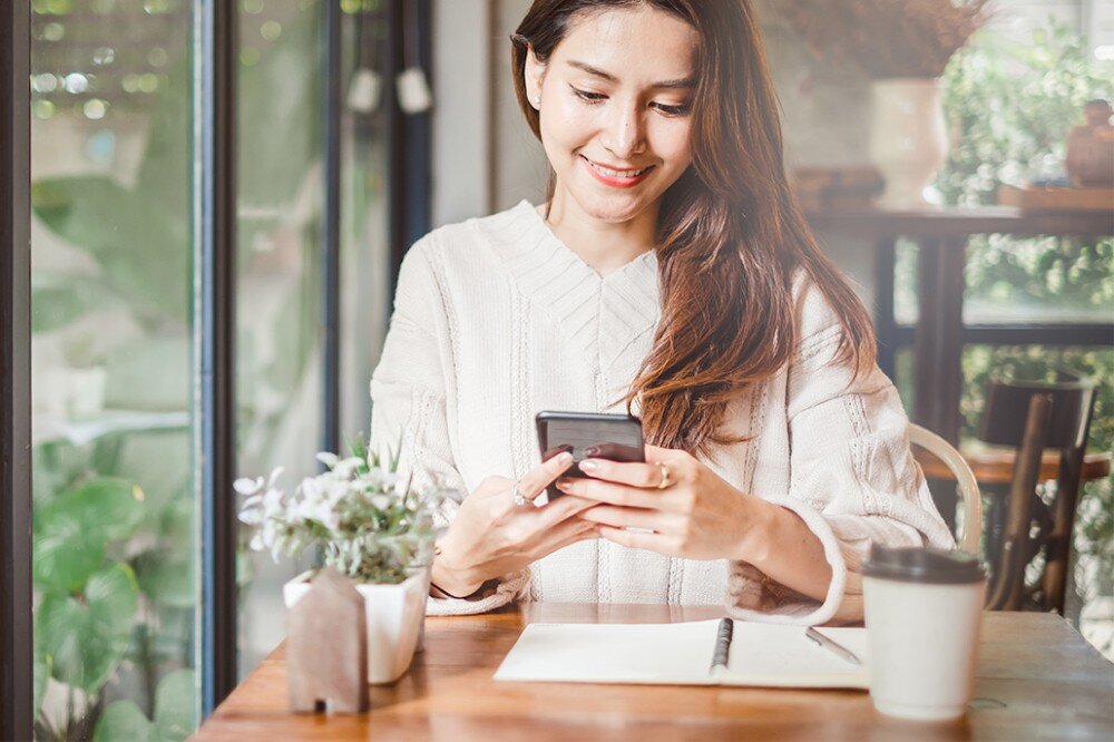 Router ASUS ZenWiFi AX Mini XD4 Plus Kobieta siedzi przy stoliku w kawiarni, trzyma smartfon i uśmiecha się, obok stoi kubek i notes, pasma, prędkość, technologie