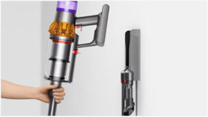 Osoba trzyma odkurzacz o metalicznym wykończeniu z fioletowym elementem na górze. Obok na ścianie zamontowany jest uchwyt z dodatkową końcówką do odkurzacza.