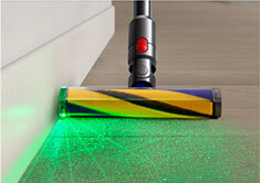 Głowica odkurzacza z żółto-niebieską szczotką, emitująca zielone światło na podłogę. Światło podkreśla drobinki kurzu na drewnianej powierzchni.