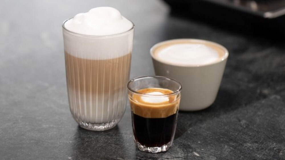 Na blacie znajdują się trzy filiżanki z różnymi rodzajami kawy: latte macchiato w wysokiej szklance, cappuccino w białej filiżance i espresso w małej szklance.