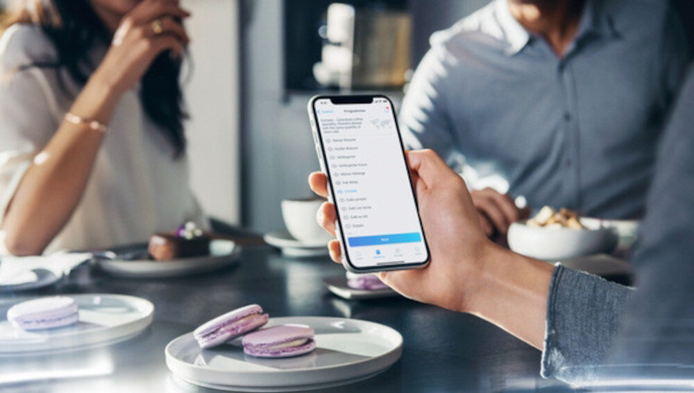 Osoba trzyma smartfon z wyświetlonym menu aplikacji, w tle widać dwie osoby przy stole z filiżankami i talerzami z makaronikami.