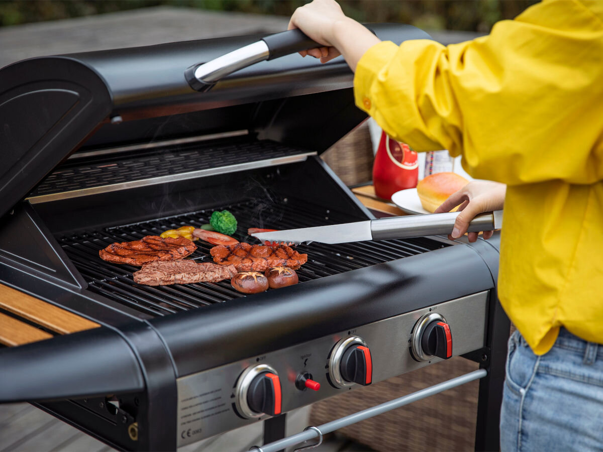 Grill gazowy YATO YG-20009 9 KW 60 x 42 cm, w tle ogród, na pierwszym planie osoba podnosi pokrywę grilla i poprawia mięso na ruszcie grilla